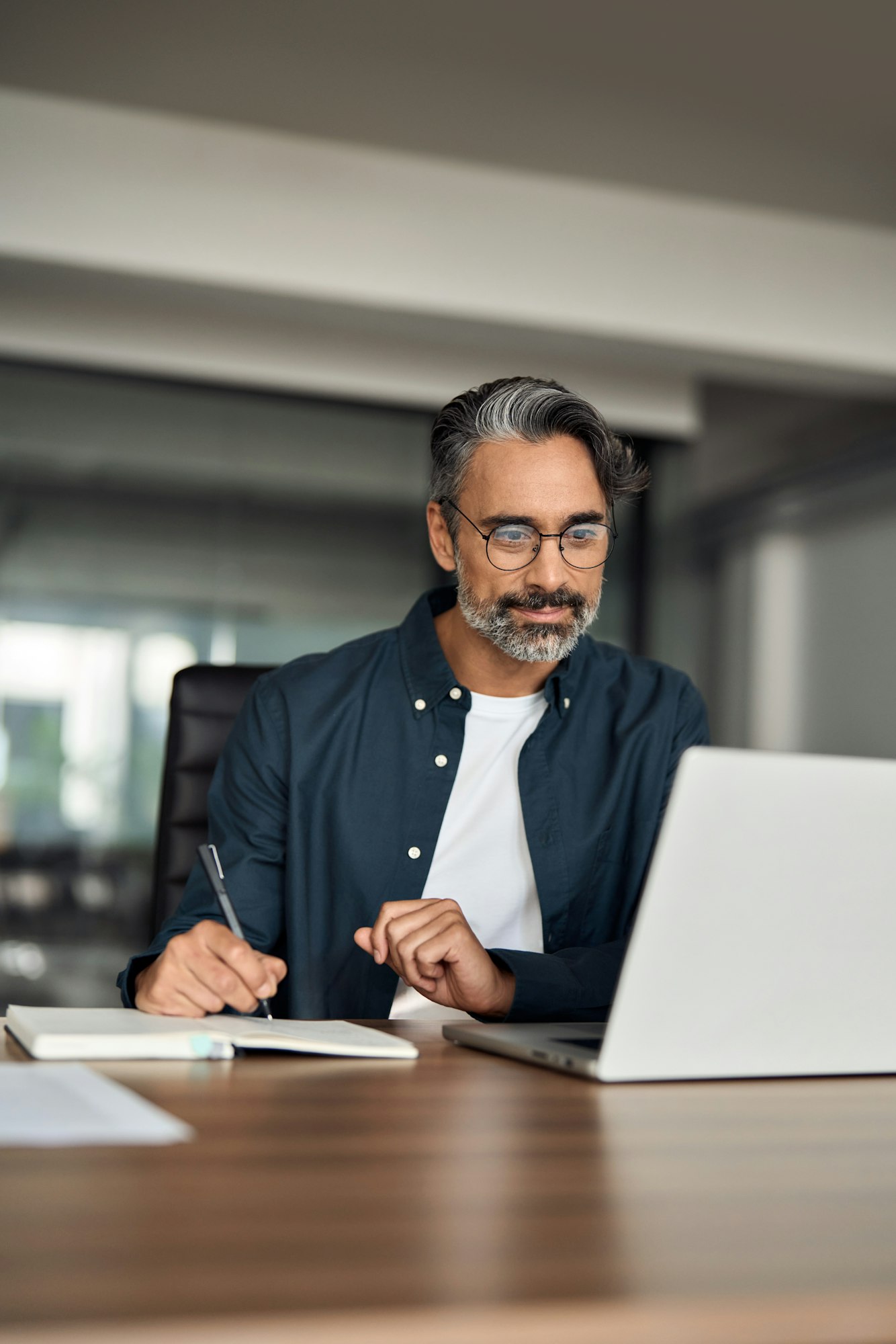 Older busy businessman writing notes learning online sitting at office desk.